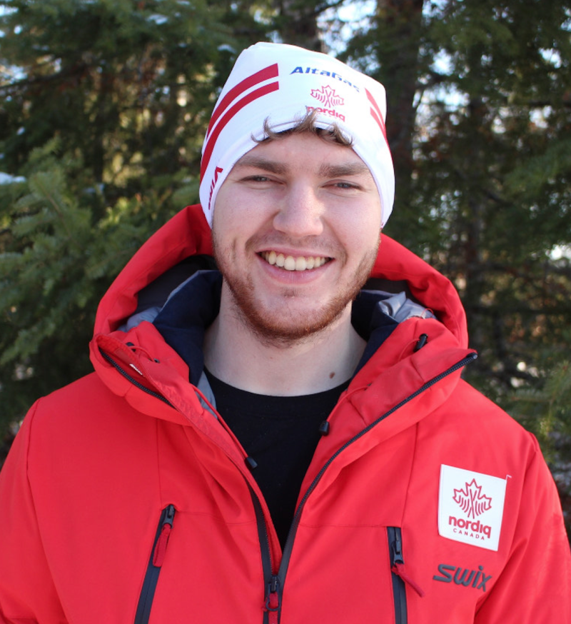 A smiling Jesse Bachinsky poses for a photo. He wears a vibrant red ski jacket, black t-shirt and a white toque.