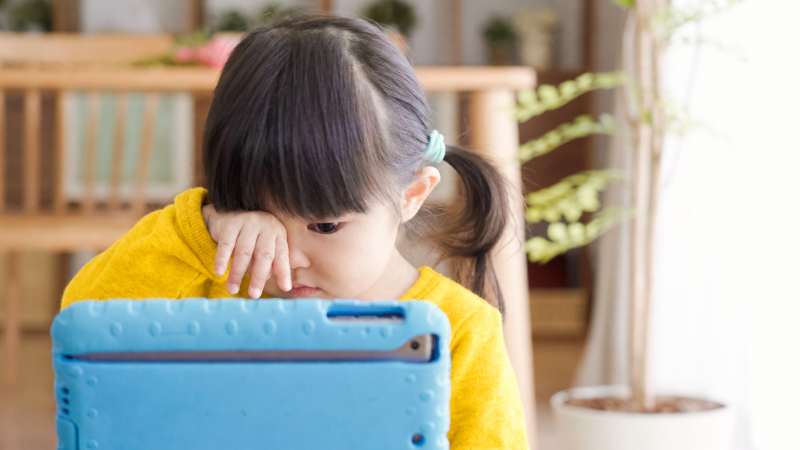 A child with dark hair rubs her eyes will looking at a tablet screen