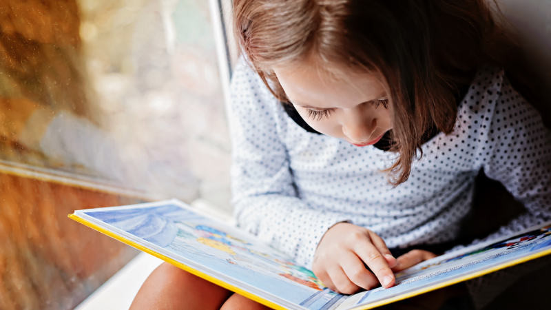 A child with brown hair reads a book while following along with her finger