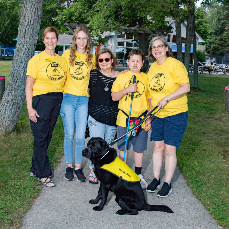 A group of five people in yellow t-shirts standing on a path with a black dog wearing a yellow vest.
