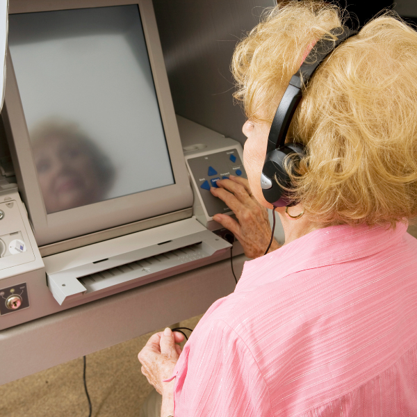 A senior woman wearing headphones uses an accessible touchscreen voting machine, with her hand on the tactile controls beside the screen.