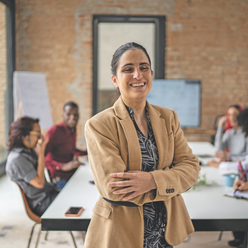 A professionally dressed woman with sight loss stands in the middle of a board room with her arms crossed. 