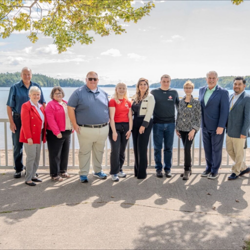 ten men and women stand in a line in front of a white picket fence. Lake Joseph is in the background. There is a blue sky.