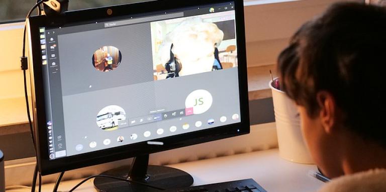 A young child sits at a desktop computer. The computer screen displays a Zoom call.