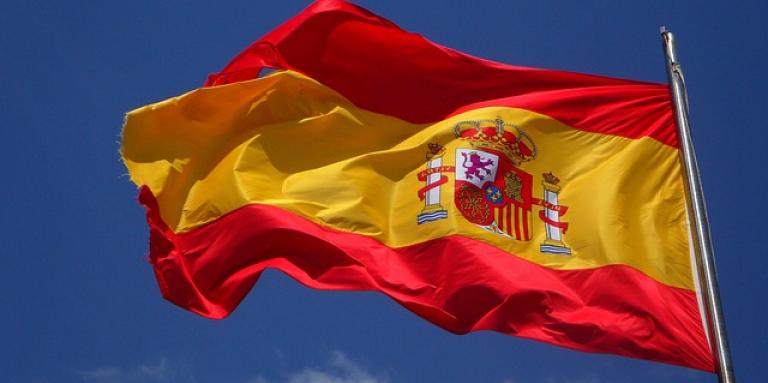 The flag of Spain on a flag post waving in the cloudy blue sky. 