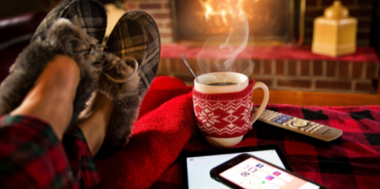 Pieds d'une personne confortablement assise devant un feu de foyer, avec une tasse chaude, des pantoufles et un téléphone intelligent.
