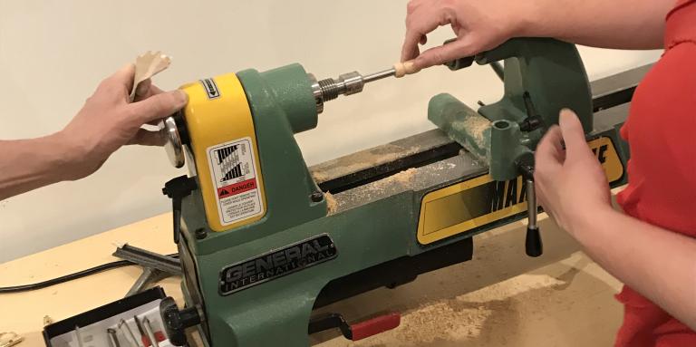 A lathe sits on a counter. Two hands maneuver a piece of wood.  
