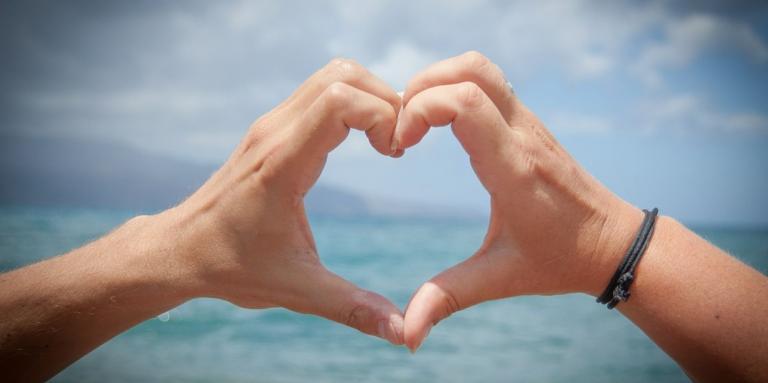 Two hands with fingers touching to create a heart shape. There is a blue sky and water in the background.