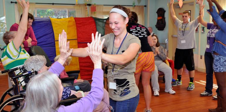A woman is standing in a circle with seniors with their hands up. She is smiling.