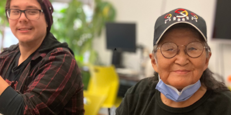 Two Indigenous participants sit at a table in CNIB Thunder Bay smiling. During a holiday program, they create decorations out of fresh cedar bows, dried orange slices, homemade gingerbread cookies, ribbons, bells, cinnamon sticks, and jute twine.