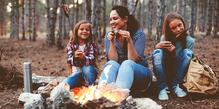A woman and two girls sit outside in front of a fire eating treats.