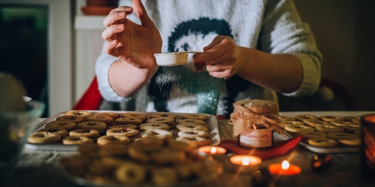 Woman sitting in front of a tray of baked goods. There are candles burning. She is sprinkling something on top.