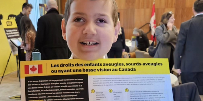 A boy is standing in a meeting room with business people behind him. He is holding up the Rights of Children Who Are blind in Canada (French version).