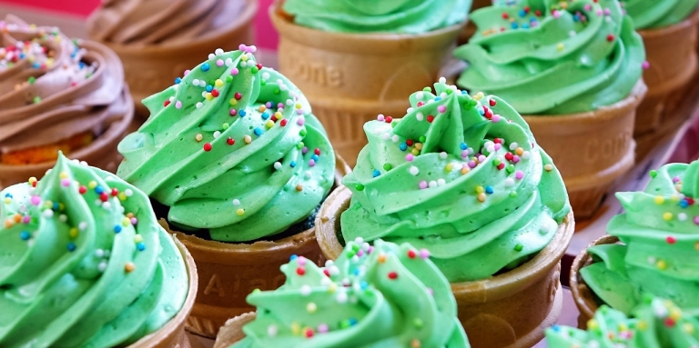 A stack of colourful cupcakes line a bake sale table. The cupcakes have green and brown icing with sprinkles. 
