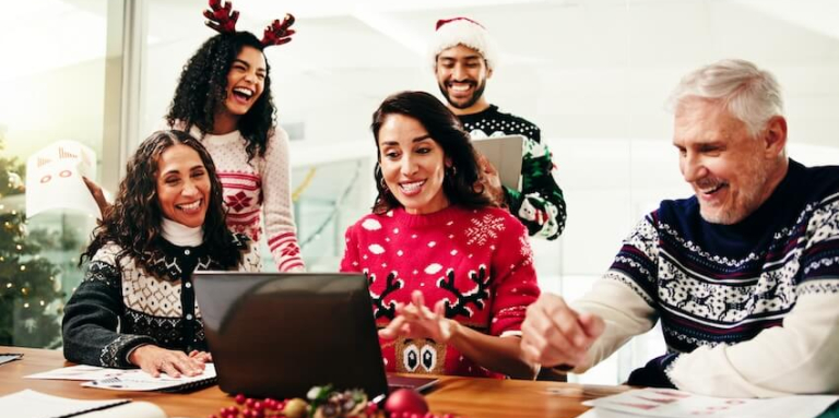 Four men and women wearing holiday sweaters and Santa hats sit around a laptop.