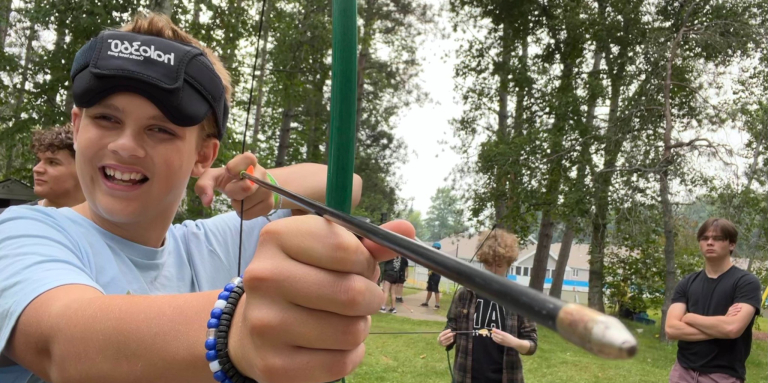 Young camper with blindfold is drawing a bow and arrow.