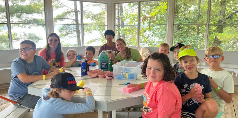 Children sitting in a gazebo at a wooden table covered with craft supplies.