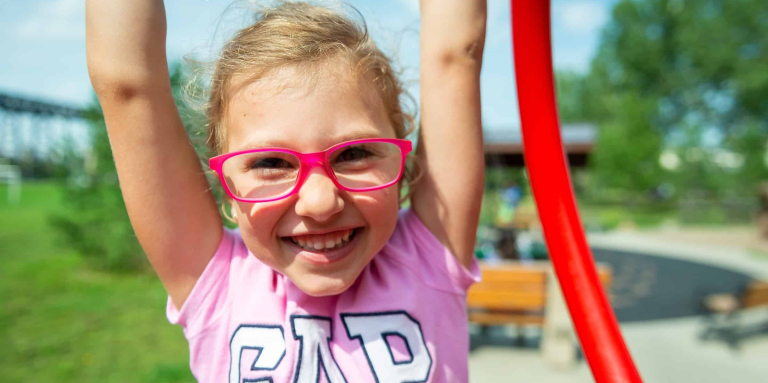Girl wearing red glasses and pink shirt is smiling and holding her arms over her head.