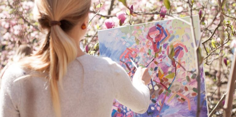 A woman is standing outside amongst bushes with blossoms. She is painting on a canvas.