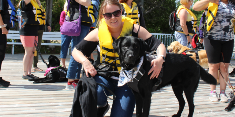A woman is kneeling on a dock with her arm around a black lab. There are people behind her with dogs. 