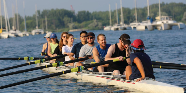 A group of eight people sit in a long rowing shell on calm water, holding oars in position. They appear relaxed and smiling, as if pausing between strokes. In the background, several sailboats are anchored near a tree-lined shore.