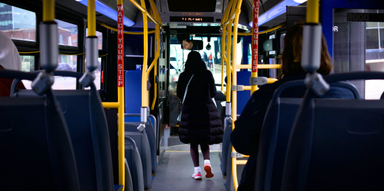 The interior of a city bus. The image is taken from the back of the bus, where a person is walking down the aisle towards the front door.
