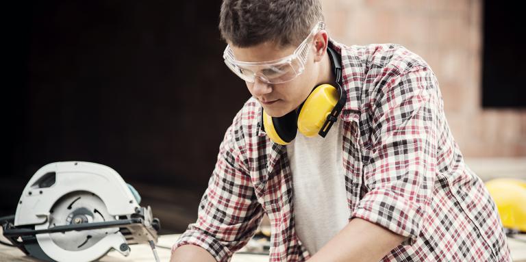 Man looking down and marking a piece of wood with a pencil. He is wearing protective glasses.