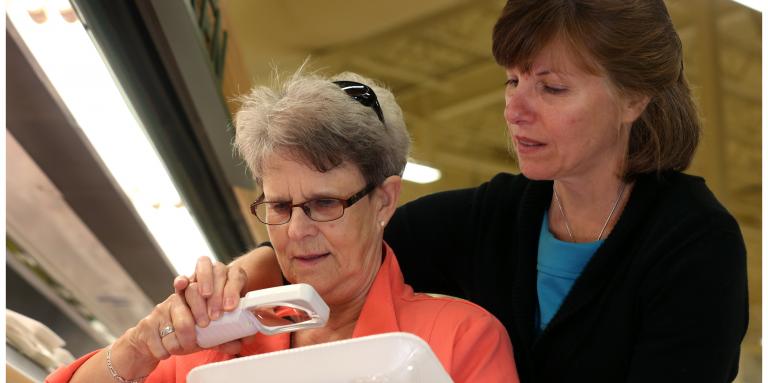 Older female holds a magnifier to a package. She is standing in front of another woman who is helping her with the magnifier.