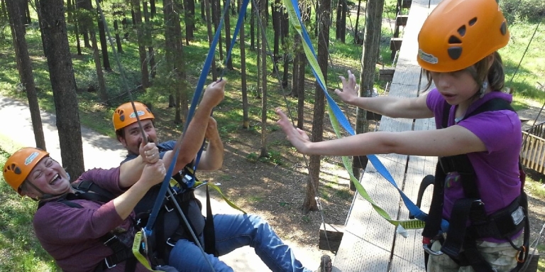 A man and a woman are leaning back attached to harnesses with their feet braced against a ziplining platform. Their daughter stands on the platform harnessed with her arms reaching toward them