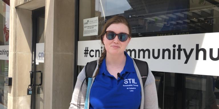 Hillary Scanlon stands in front of a CNIB community hub wearing the shirt with her brand on it: Sustainability Through an Inclusive Lens.