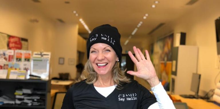 Denise Justin smiles and waves, wearing a shirt and toque with the Say Hello logo. She's standing in a CNIB hub in Toronto.