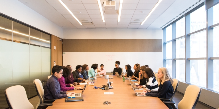 A group of about twenty people sit around a long conference table working on computers and looking at a speaker