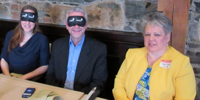 Cindy sits with two Dining in the Dark guests, who are wearing blindfolds.