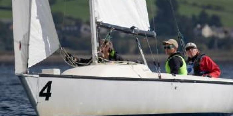 oshua Cook et deux autres personnes sont assises dans un bateau à voile sur l'eau. Ils sont aux championnats du monde de voile aveugle en Ecosse.