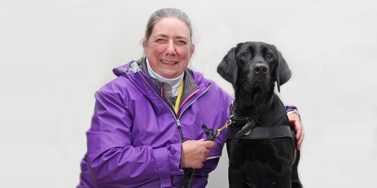 A woman in a purple jacket and her black Lab/Golden Retriever cross guide dog.