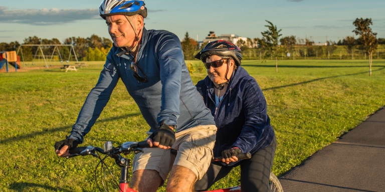 A man and woman riding a red tandem bike through the park on a sunny day.