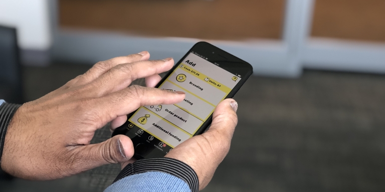 Image of a set of hands holding an iPhone with the Venture Zone Game app open on the screen, displaying the game’s “Add” menu, which lets players choose between various branding, marketing, production or funding options. 