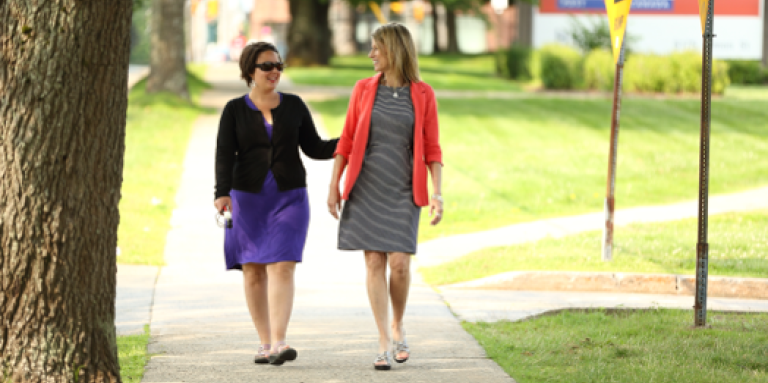 An example of the sighted guide technique. Two woman walk down a sidewalk together. A woman holds the guide's arm lightly above the elbow and allows the guide to walk one-half step ahead.