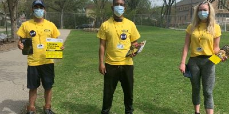 Three fundraisers in yellow CNIB T-shirts, lanyards and protective face masks stand apart on a grassy field, holding up CNIB fundraising brochures. 