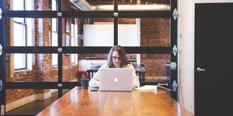 A young woman sits alone in a boardroom. She is busy typing on her laptop