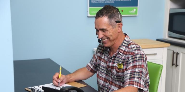 A smiling man wearing a telephone headset takes notes on a clipboard.