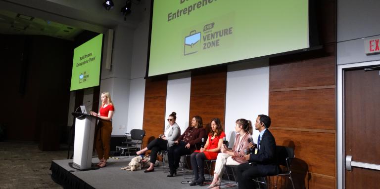 A group of panelists sits on stage at Connecting the Dots. Behind them is a slideshow screen which displays "Bold Dreams - Entrepreneur Panel."