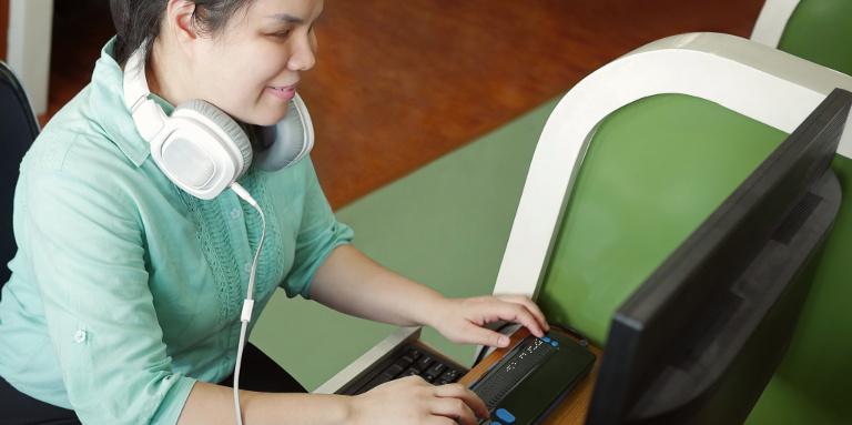 A young woman with headphones on sits at her desktop computer and types on a refreshable a braille display.