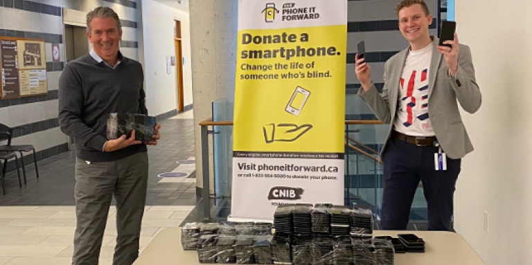 A smiling John M. Rafferty & Thomas Simpson stand at a table and hold multiple donated smartphones from the Bank of Canada. The table is also stacked with donated smartphones. Behind them stands a Phone it Forward banner display.