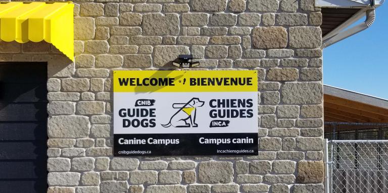 Une photo du panneau de bienvenue placé à l'extérieur du Campus canin. On peut y lire : « Welcome et Bienvenue » avec le logo des chiens-guides d'INCA au milieu du panneau