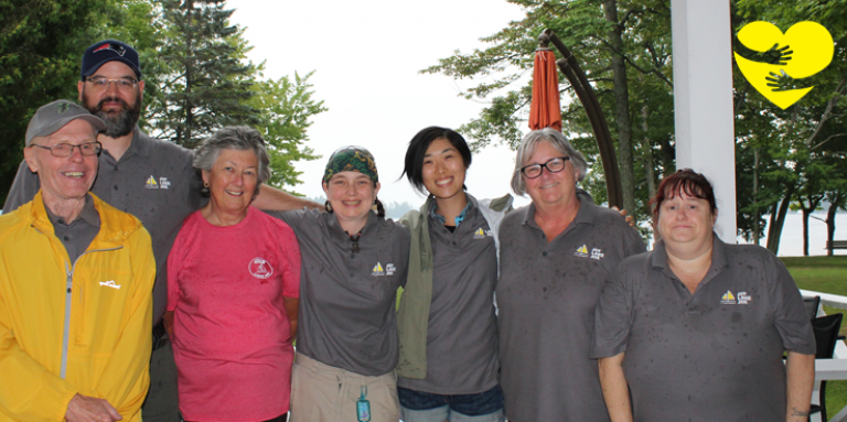 Sandra, portant une chemise rose, sourit pour la photo de groupe avec d'autres bénévoles d'INCA au camp Lake Joe. Un dessin de bras serrant un cœur jaune illustré se trouve dans le coin supérieur gauche de la photo.
