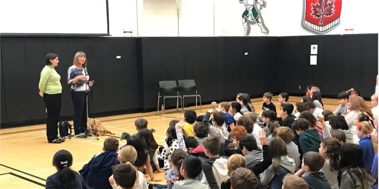 A CNIB Ambassador Presentation in front of a large group of school-aged children sitting in a gym.