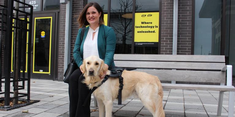 A smiling woman sitting on a bench with her CNIB Guide Dog, outside a CNIB building with yellow branded CNIB signs and posters. 
