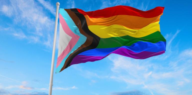 A Progress Pride flag waves in a cloudy sky.