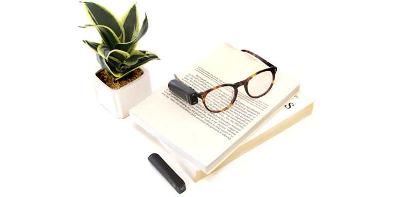 Orcam sur des lunettes déposées sur un livre. Une plante est à gauche du livre.